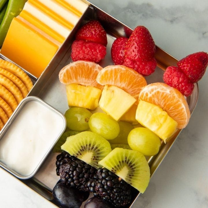 Rainbow fruit skewers in a metal lunch container with yogurt dip, cheese, crackers, and veggies.