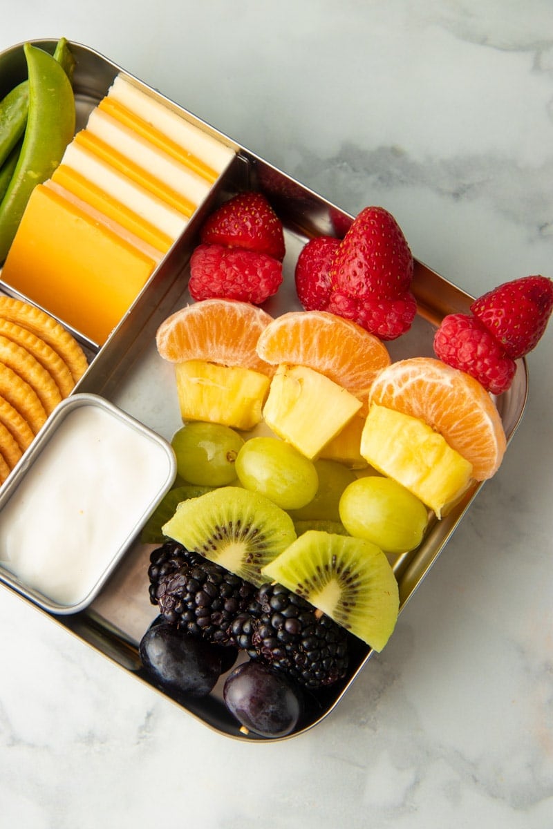 Rainbow fruit skewers in a metal lunch container with yogurt dip, cheese, crackers, and veggies.