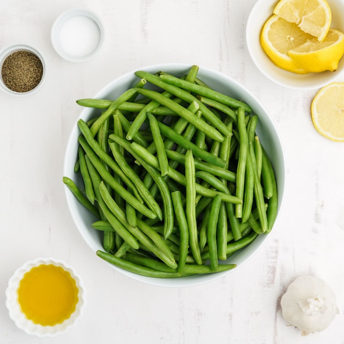 Overhead view of raw green beans, olive oil, lemon, garlic, salt, and pepper in small bowls