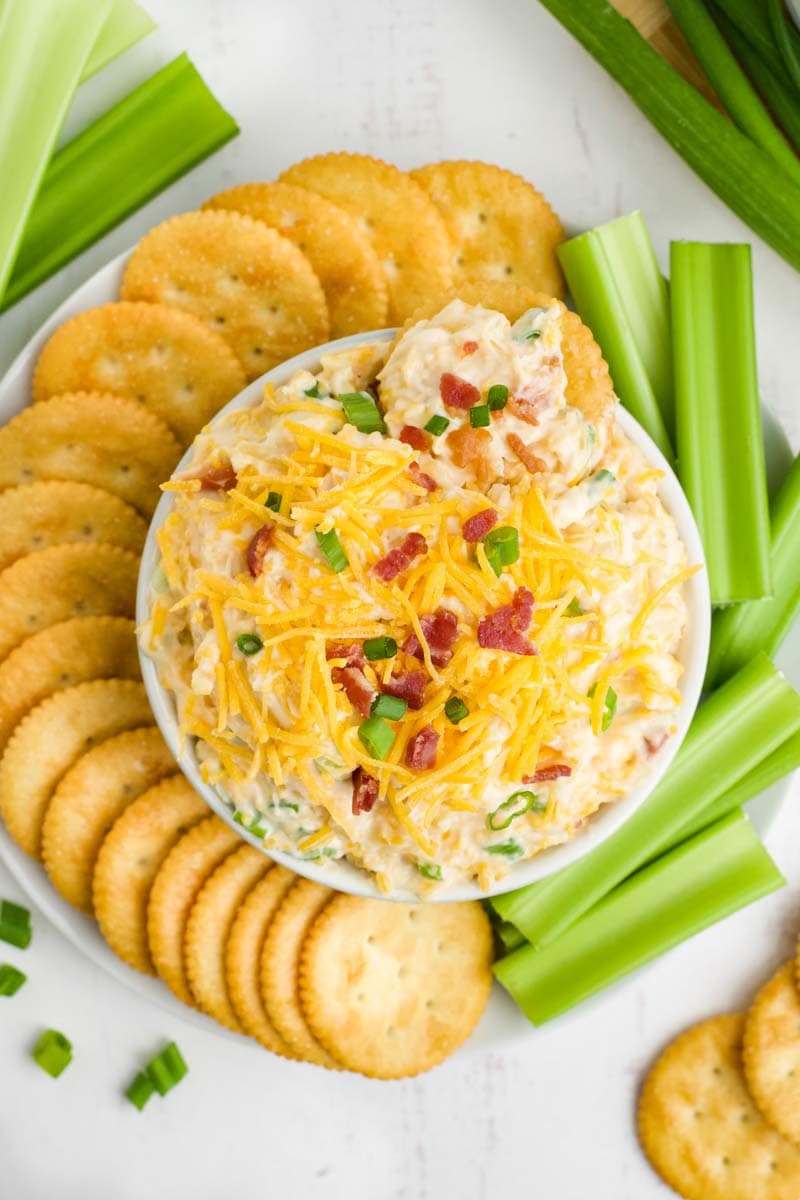 Overhead of million dollar dip in a large serving ramekin surrounded by round crackers and celery sticks on an appetizer platter.