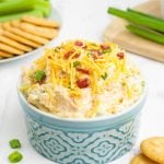 A large serving ramekin of creamy million dollar dip stands on a counter with crackers and celery sticks plated nearby.