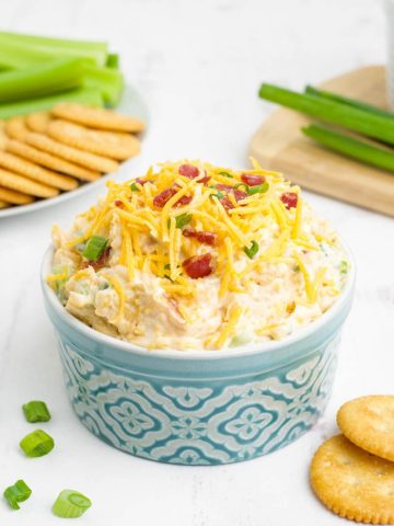 A large serving ramekin of creamy million dollar dip stands on a counter with crackers and celery sticks plated nearby.