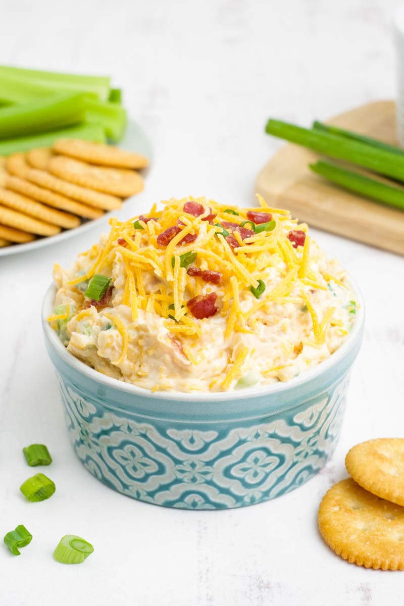 A large serving ramekin of creamy million dollar dip stands on a counter with crackers and celery sticks plated nearby.
