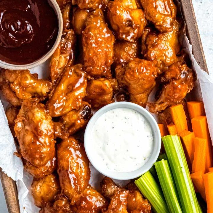 Overhead of a wooden serving tray lined with parchment paper filled with baked wings, bowls of barbecue sauce and ranch dip, and celery and carrot sticks.