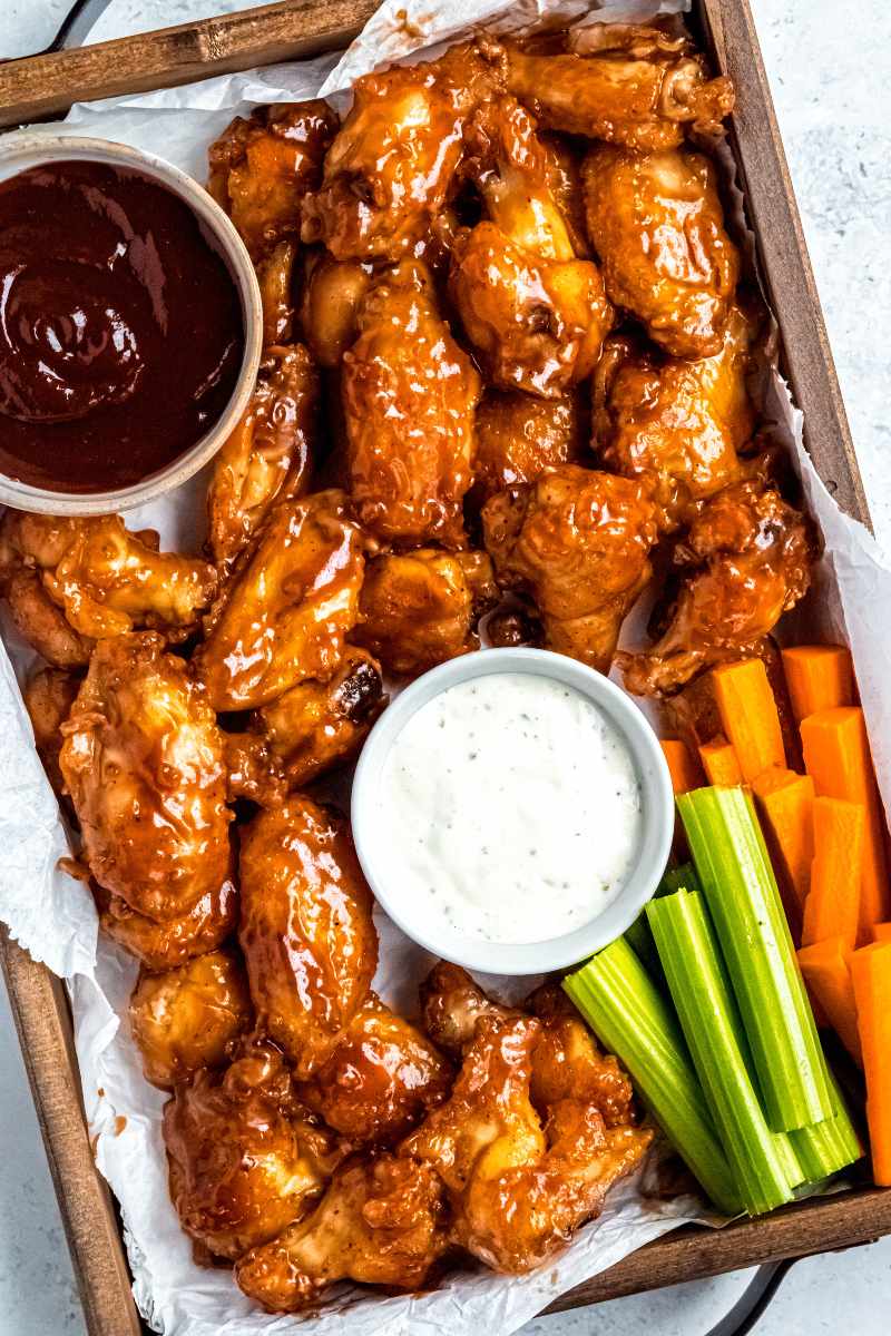 Overhead of a wooden serving tray lined with parchment paper filled with baked wings, bowls of barbecue sauce and ranch dip, and celery and carrot sticks.