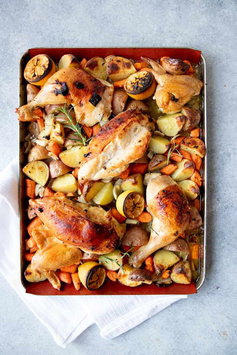 Overhead of a sheet pan loaded with roasted root veggies topped with roast chicken pieces and lemon halves.