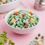 A small white bowl of green pea salad sits on a light pink counter with fresh green parsley nearby.