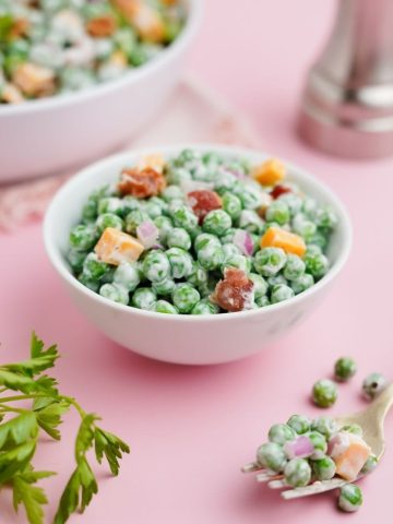 A small white bowl of green pea salad sits on a light pink counter with fresh green parsley nearby.