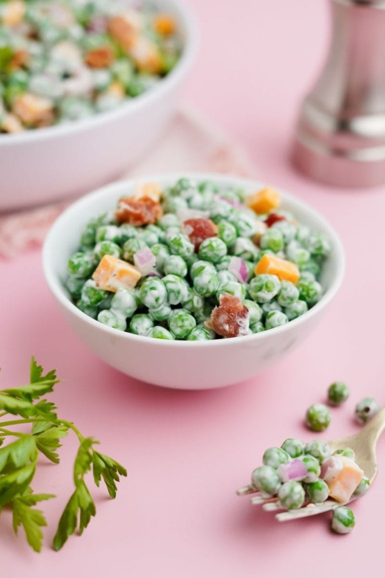 A small white bowl of green pea salad sits on a light pink counter with fresh green parsley nearby.