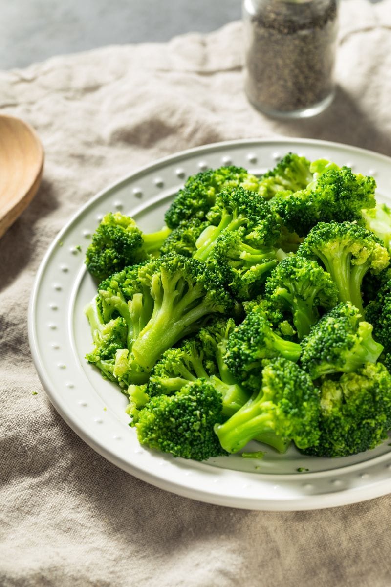 A serving platter of steamed broccoli on a light kitchen linen additional black pepper nearby.