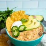 A teal bowl filled with Instant Pot white chili topped with sour cream, avocado, jalapeno, and corn tortilla chips.