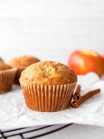 A perfectly domed apple cinnamon muffin rests on a parchment-lined cooling rack with two cinnamon sticks beside it.