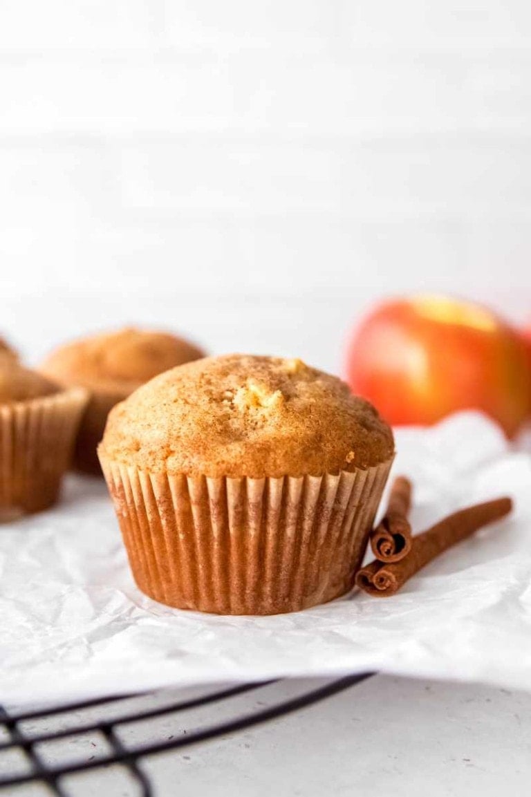 A perfectly domed apple cinnamon muffin rests on a parchment-lined cooling rack with two cinnamon sticks beside it.
