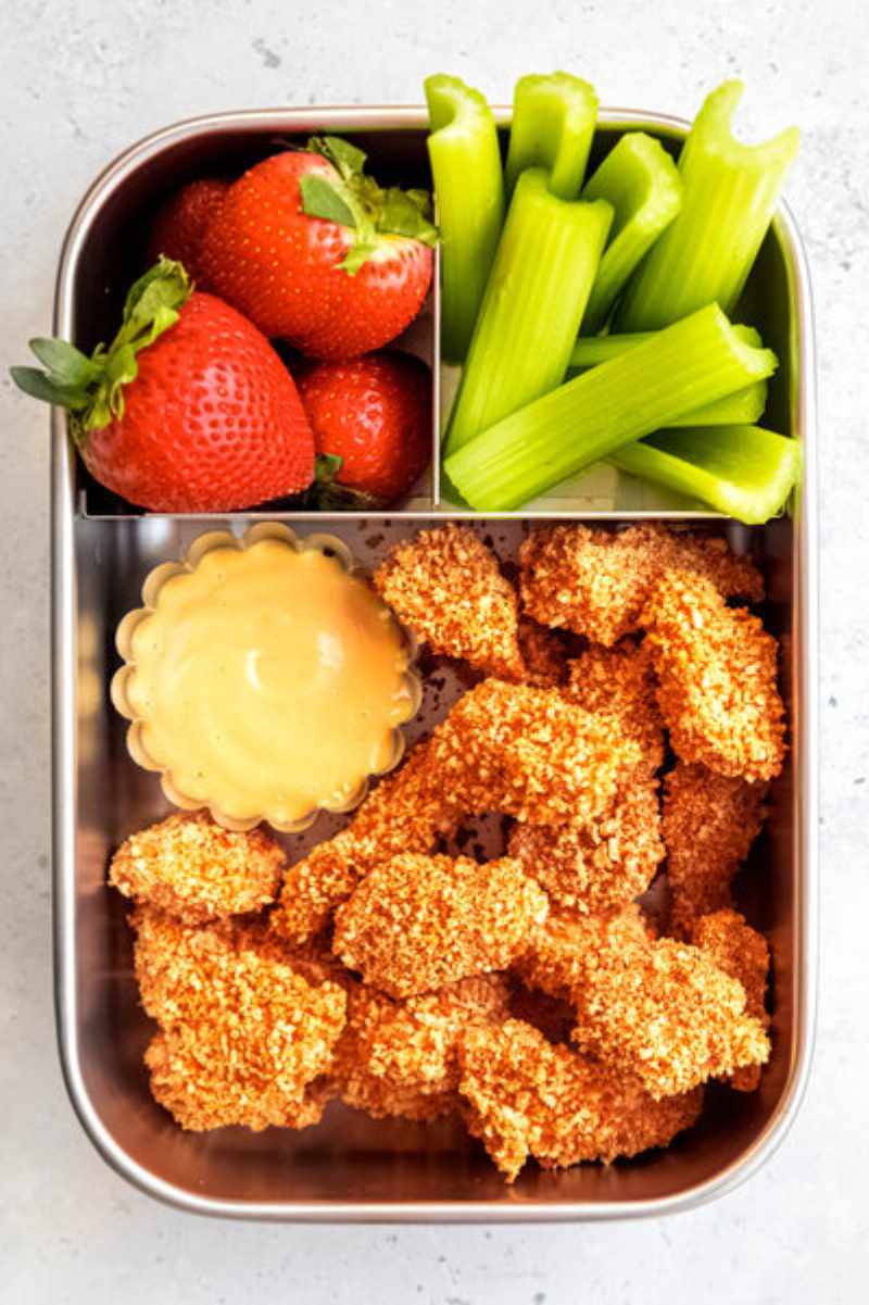 A stainless steel bento-style lunch box filled with homemade chicken nuggets, strawberries, and celery sticks.