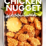 A stainless steel bento-style lunch box filled with homemade chicken nuggets, strawberries, and celery sticks. A text overlay reads, "Homemade Chicken Nugget School Lunch."