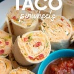 Close view of taco pinwheels piled on a white plate around a small teal bowl of salsa for dipping. A text overlay reads, "Chicken Taco Pinwheels."