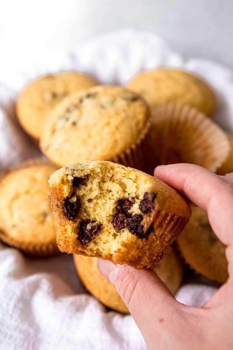 A hand holds a chocolate chip muffin with a bite out of it showing the tender, chocolate-speckled center.