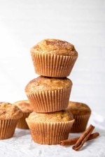 Three cinnamon sugar muffins stacked on a white background with additional muffins and cinnamon sticks alongside.