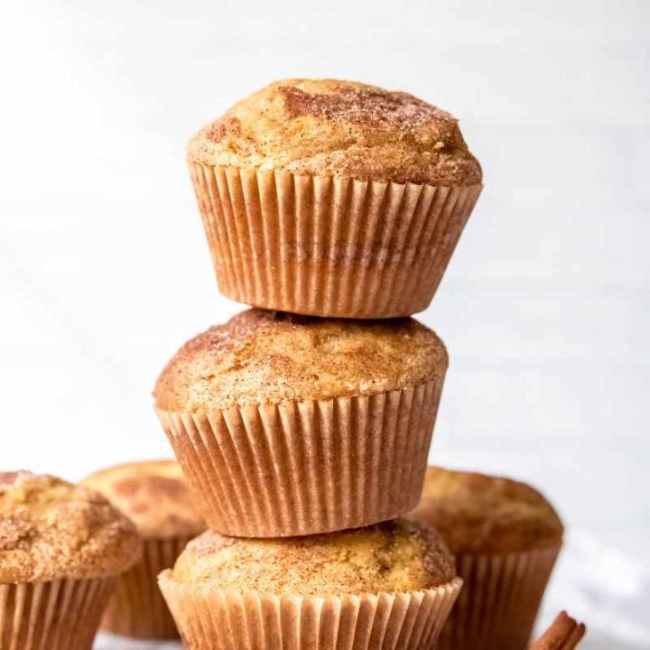Three cinnamon sugar muffins stacked on a white background with additional muffins and cinnamon sticks alongside.