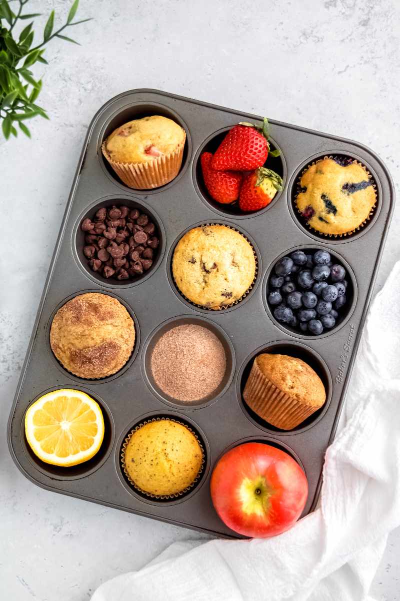 A muffin tin filled in alternating cups with six flavors of muffins and their corresponding mix-ins, such as strawberries, chocolate chips, blueberries, and more.