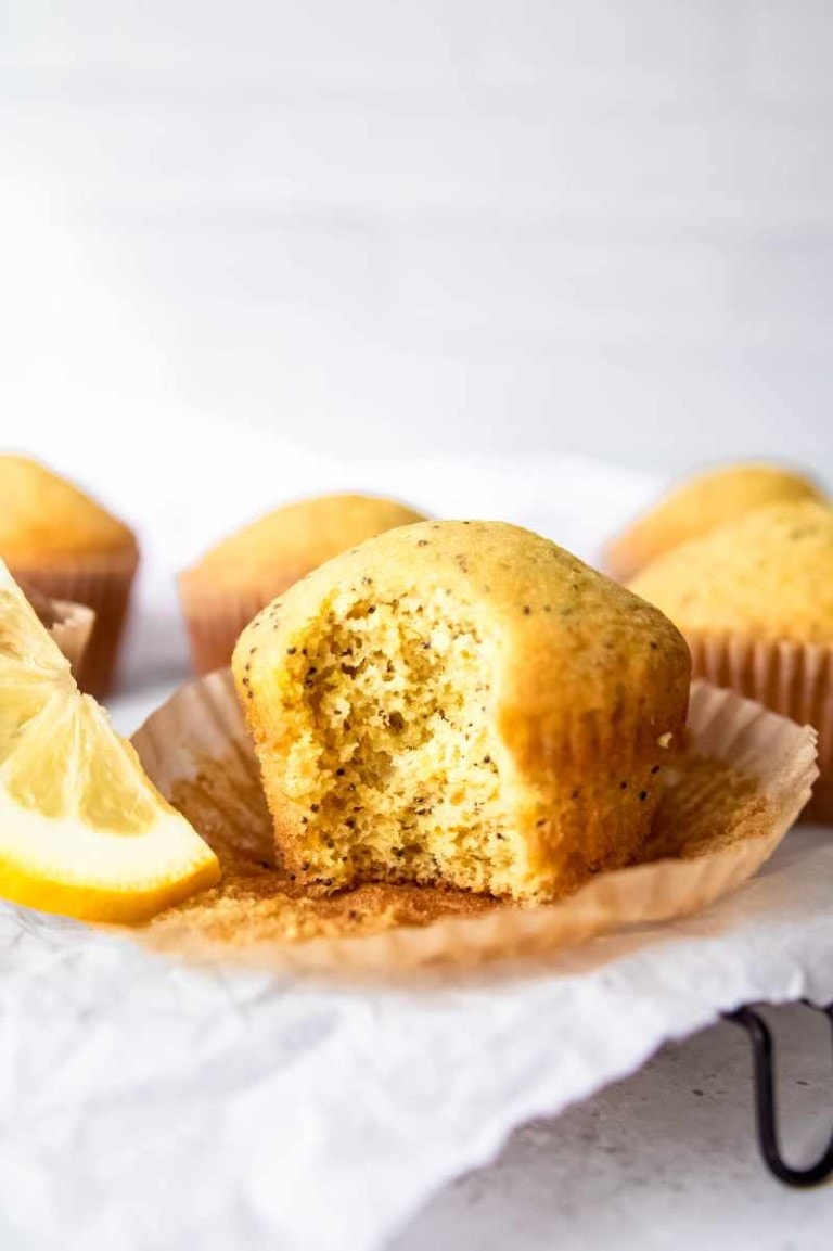 A lemon poppy seed muffin with a bite taken out, showing the speckled inside, sits in its peeled liner on a light background.