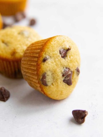 A mini muffin with chocolate lays on its side on a light countertop with a chocolate morsel nearby.