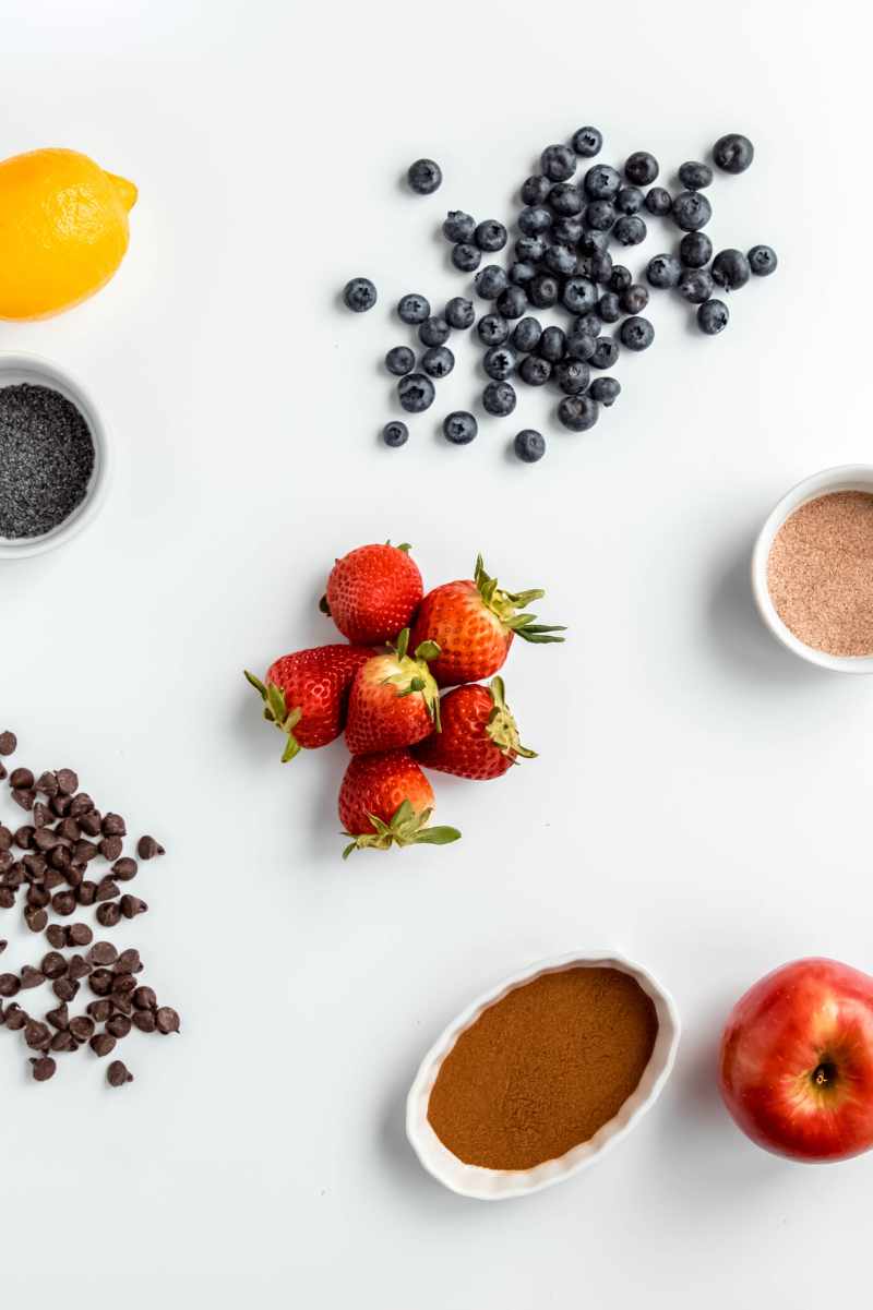 Overhead of flavorful mix-ins to stir into a muffin batter, including fresh blueberries and strawberries, chocolate chips, cinnamon, poppyseeds and lemon.
