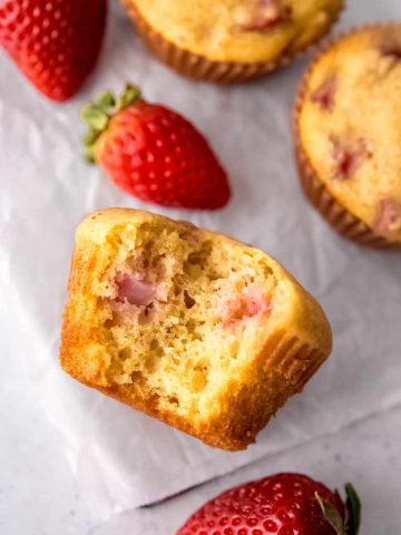 Close view of a strawberry muffin lying on its side with a bite taken out of it showing pockets of cooked pink fruit inside.