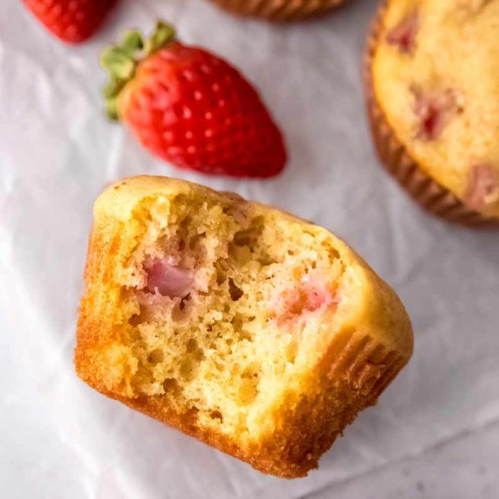Close view of a strawberry muffin lying on its side with a bite taken out of it showing pockets of cooked pink fruit inside.