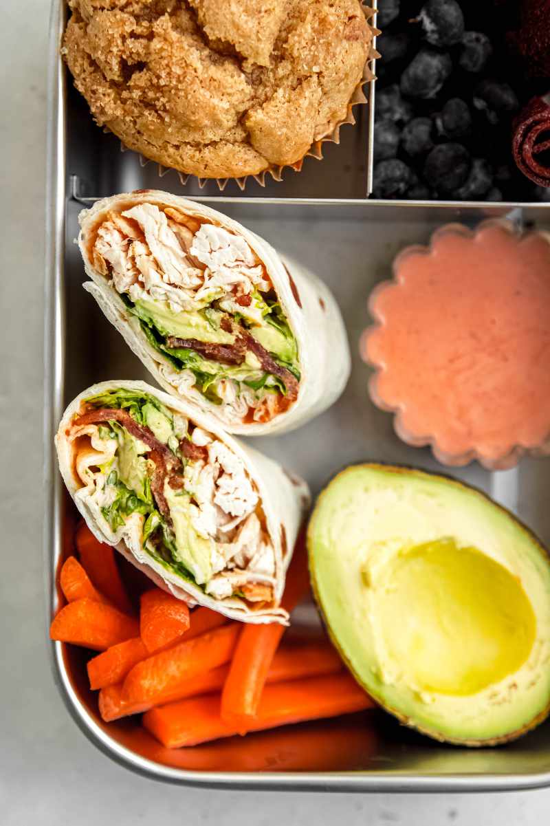 Two halves of a chicken wrap with spicy ranch dipping sauce packed in the large lunchbox compartment with carrot sticks and an avocado half.