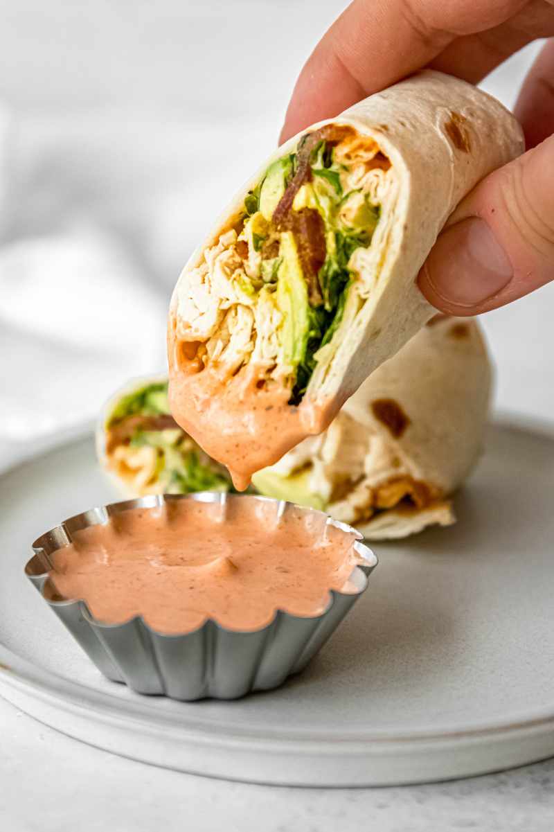 A hand dips a chicken bacon ranch wrap cut on the bias into a small scalloped bowl of spicy ranch dip.