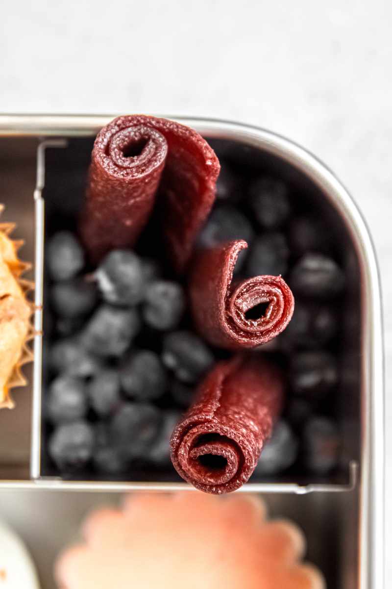 Close view of three pieces of fruit leather tucked into one section of a bento-style lunchbox with fresh blueberries.