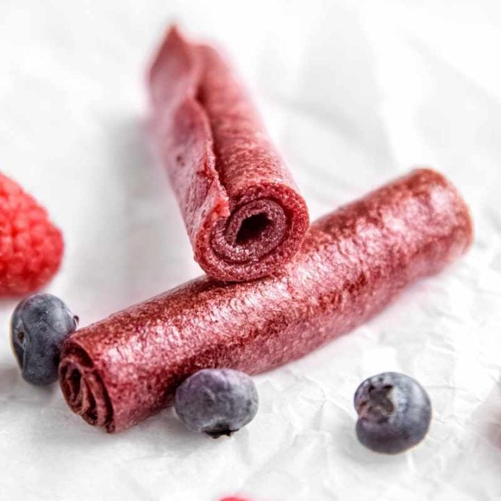 Two pieces of fruit leather rolled and stacked with the end of one on the center of the other, fresh berries around them.