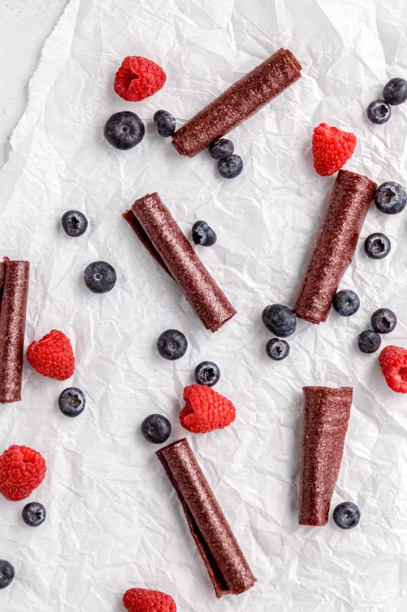 Homemade fruit leather rolls on white parchment paper with fresh raspberries and blueberries scattered around them.