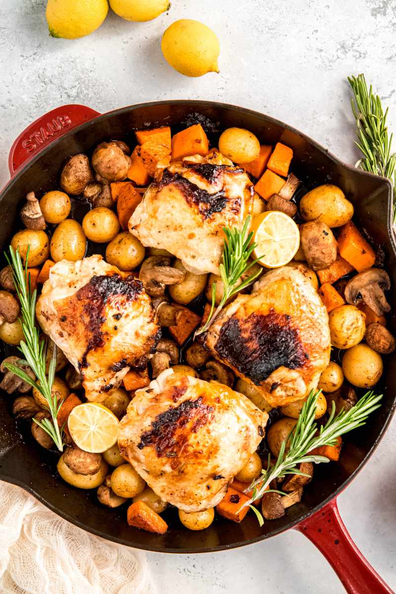 Top view of a lemon chicken skillet family dinner with four chicken thighs on top of a bed of fall veggies in an enameled cast iron skillet.