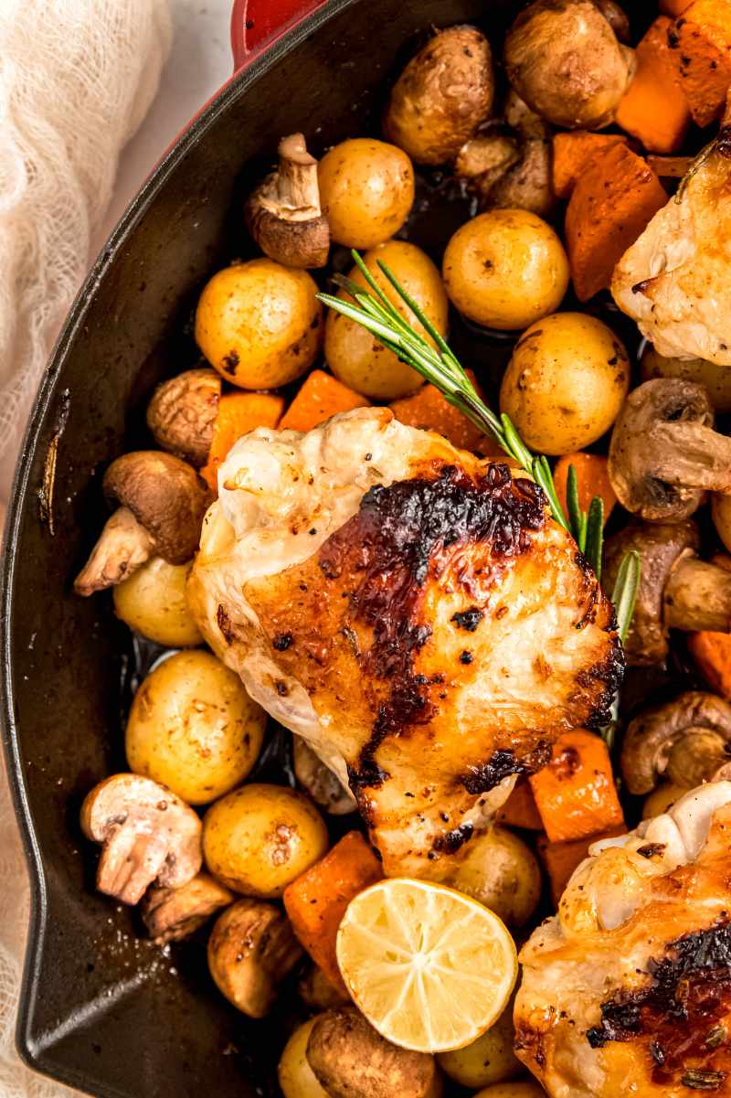 Close view of a chicken thigh with perfectly seared skin sitting atop a bed of veggies in a cast iron skillet with a sprig of rosemary and a lemon half nestled beside it.