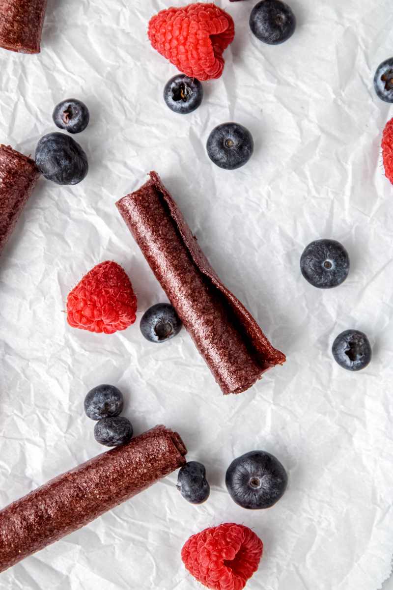 A rolled fruit snack rests on parchment paper with fresh berries around it, the end pulled slightly open as if someone started to unroll it.
