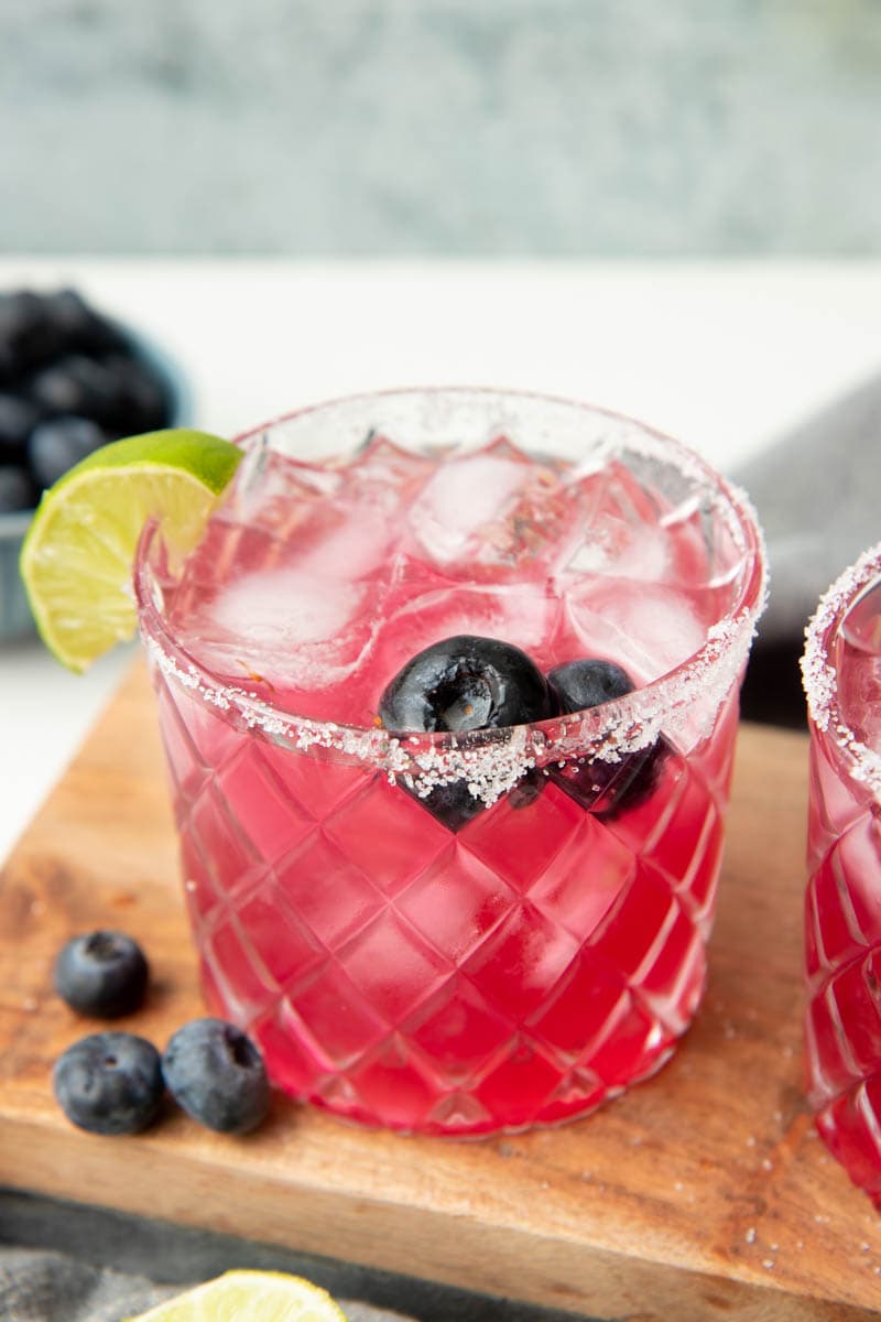 Close view of a bright pink blueberry margarita in a rocks glass with a salt rim, two blueberries, and a lime wedge garnishing the glass.