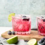 Two blueberry margaritas served on a wooden board in rocks glasses with salt rims, blueberries, and lime wedges.