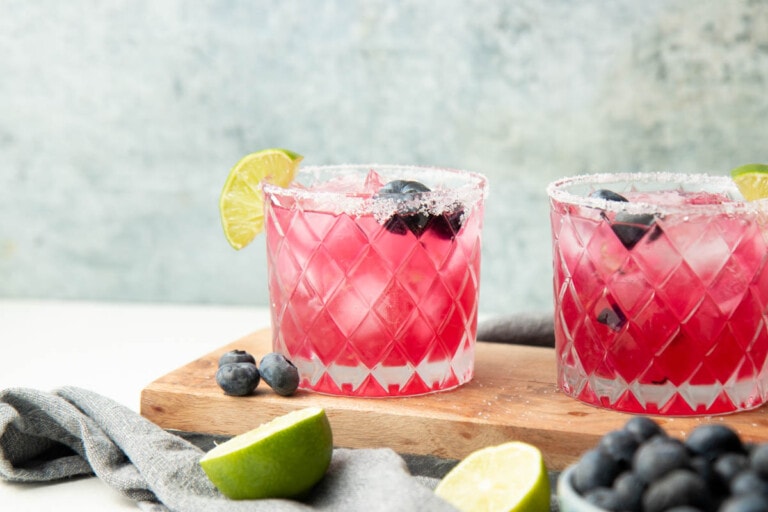 Two blueberry margaritas served on a wooden board in rocks glasses with salt rims, blueberries, and lime wedges.