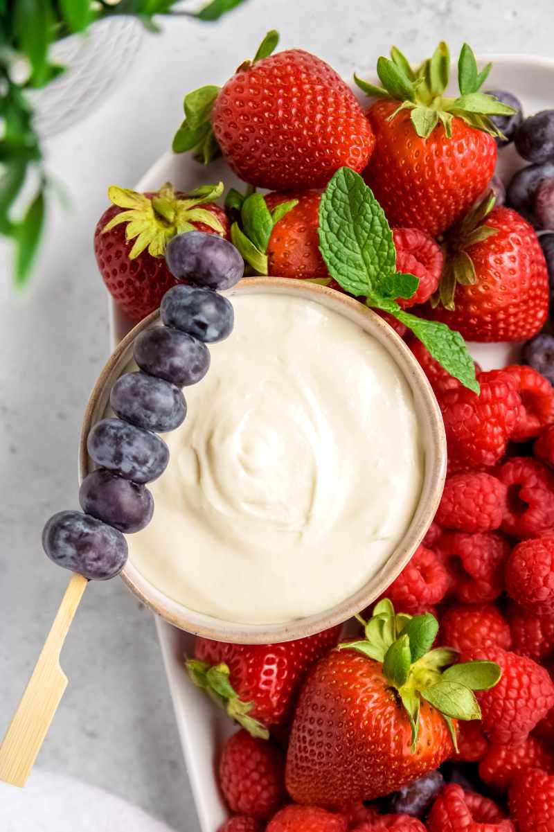 A skewer of fresh blueberries rests across the rim of a bowl of creamy dip surrounded by other fresh berries.