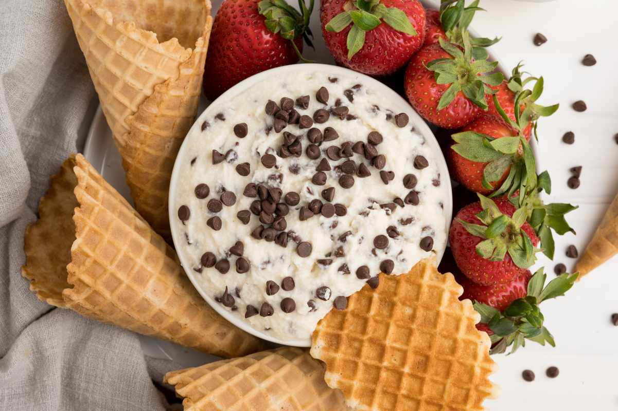 Close view of a waffle cone piece dunked into a bowl of cannoli dip, more waffle cones and fresh strawberries arranged around it.