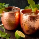 Two copper mugs side by side on a metal serving tray filled with chilled bourbon cocktails garnished with mint and lime.