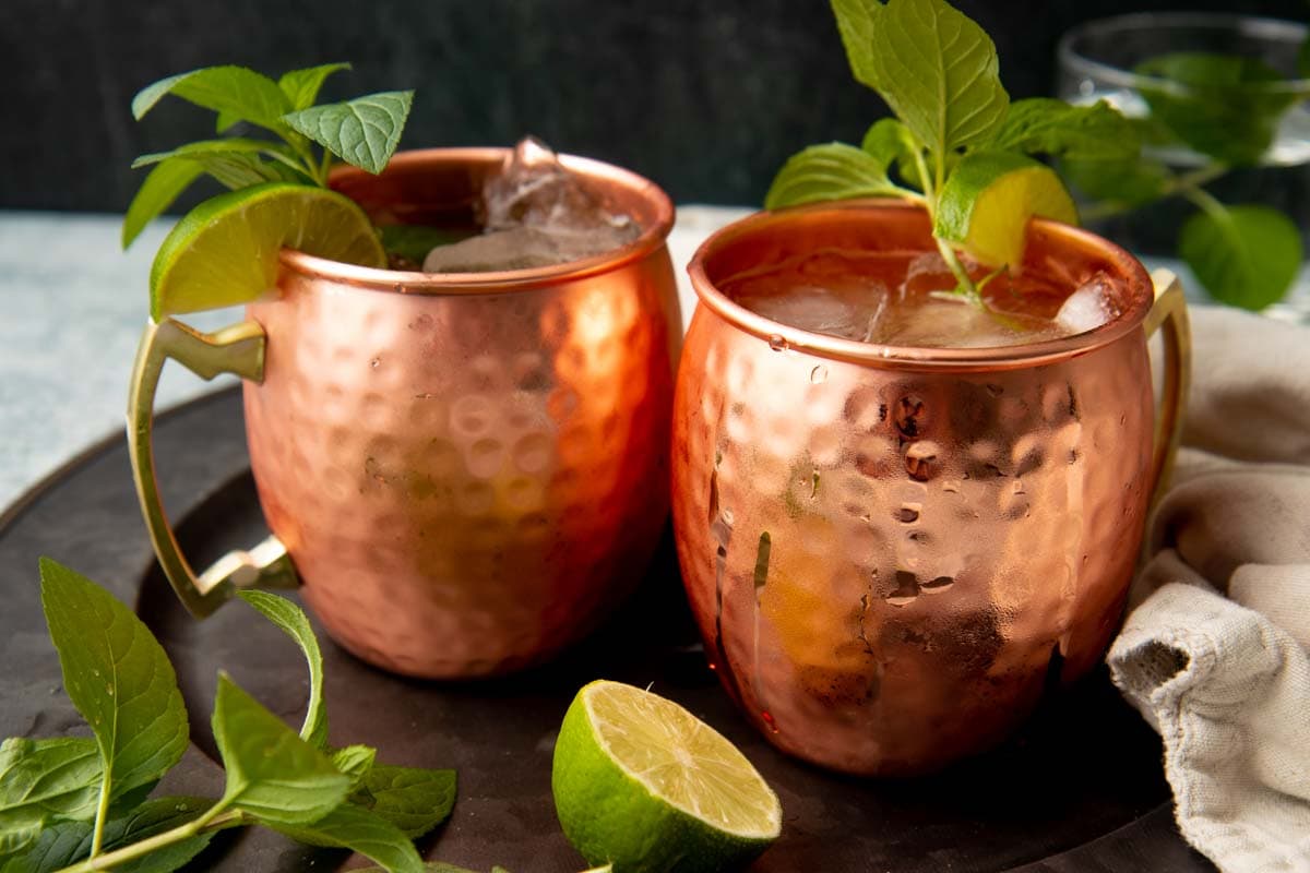 Two copper mugs side by side on a metal serving tray filled with chilled bourbon cocktails garnished with mint and lime.