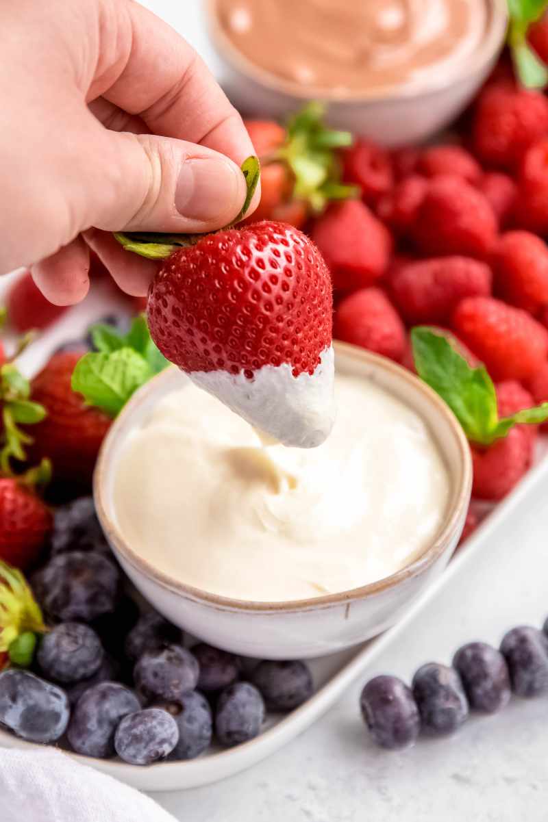 A hand dips a bright red strawberry into creamy dip.