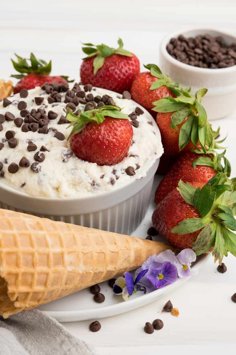 A bright red strawberry rests in a bowl of creamy dessert dip with more fresh berries and waffle cones around it.