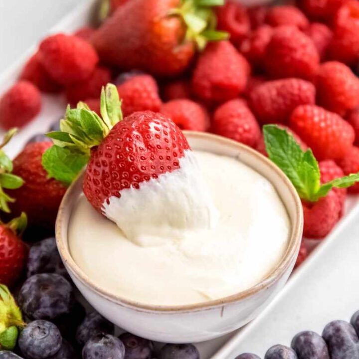 A strawberry rests in a bowl of creamy fruit dip on a serving tray with other fresh fruit.