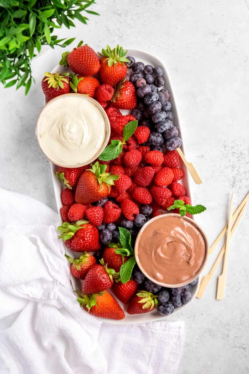 A long platter of beautiful fresh berries including strawberries, raspberries, and blueberries with two bowls of marshmallow fruit dip, one classic and one chocolate.