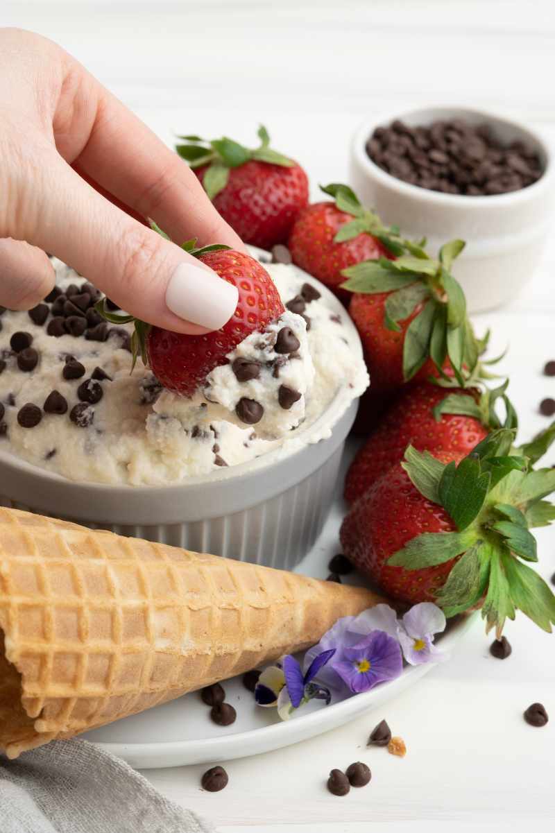 A hand dips a strawberry into a serving bowl of cannoli dip garnished with mini chocolate chips.