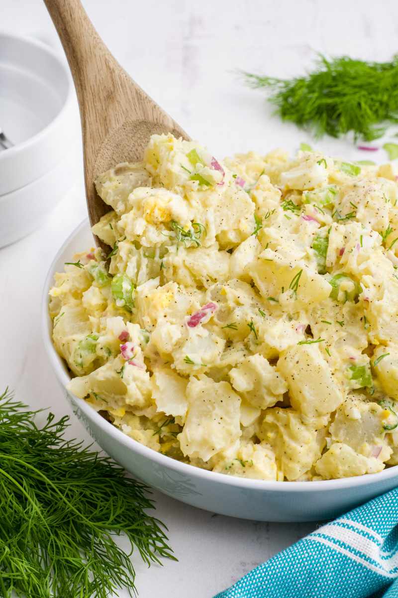 A wooden spoon scoops potato salad from a large white bowl.
